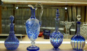 Quatre carafes en verre bleu et transparent ornées de formes et de motifs différents sont présentées sur une étagère en bois sous un éclairage vif.