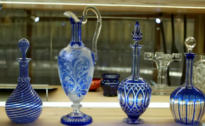 Quatre carafes en verre bleu et transparent ornées de formes et de motifs différents sont présentées sur une étagère en bois sous un éclairage vif.