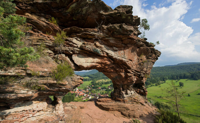 Le Rocher du Wachtfels. Une arche de grès naturelle surplombe un petit village et des champs verdoyants, avec des collines boisées et un ciel partiellement nuageux en arrière-plan.