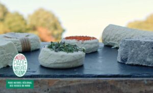 Une variété de fromages artisanaux, certains agrémentés d'herbes ou d'épices, sont présentés sur une ardoise noire en plein air, avec des arbres en arrière-plan. Le logo d'un parc régional apparaît dans un coin.