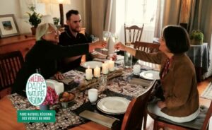 Trois personnes sont assises autour d'une table dressée pour un repas, levant leurs verres pour trinquer. La table est décorée de bougies et d'un chemin de table en dentelle.