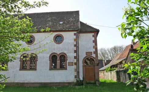 Un bâtiment blanc avec des fenêtres cintrées, une porte en bois et un toit de tuiles, entouré d'herbe et de verdure par une journée ensoleillée.
