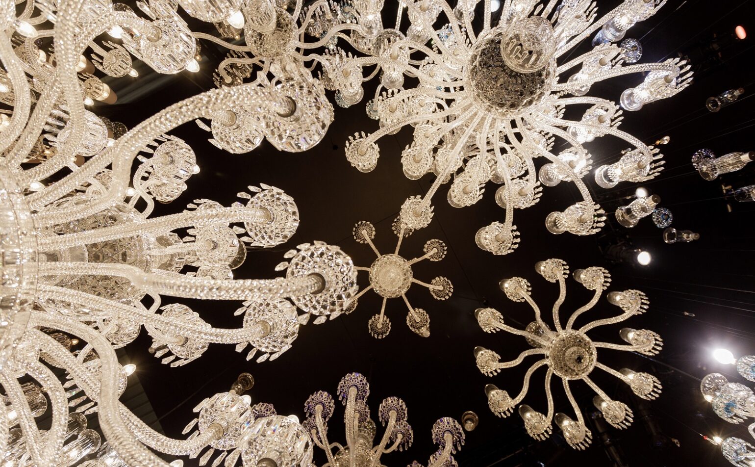Regardant vers le haut plusieurs lustres en verre ornés de motifs complexes, placés sur un plafond sombre avec de petits spots.