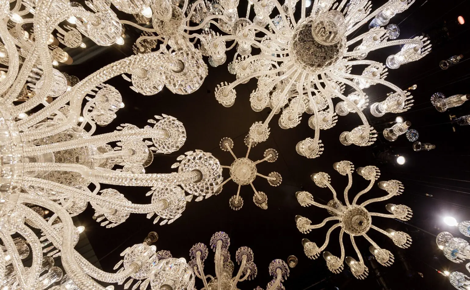 Regardant vers le haut plusieurs lustres en verre ornés de motifs complexes, placés sur un plafond sombre avec de petits spots.