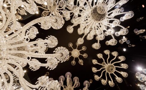 Regardant vers le haut plusieurs lustres en verre ornés de motifs complexes, placés sur un plafond sombre avec de petits spots.