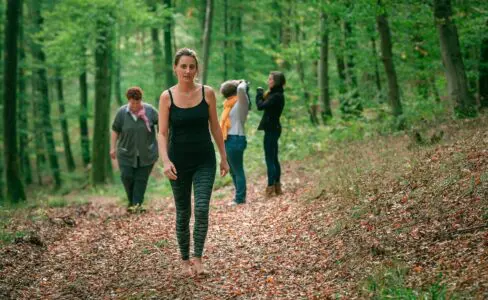 Quatre adultes marchent dans une zone boisée pendant la journée ; une femme marche pieds nus au premier plan, tandis que trois autres sont à l'arrière-plan.