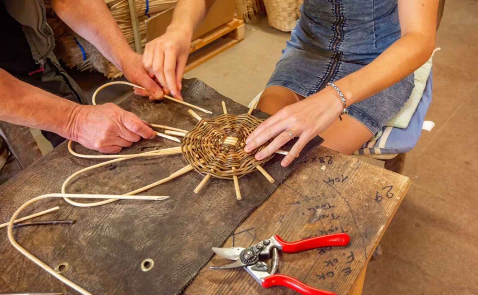 Deux personnes tissent un panier circulaire en osier sur un établi, avec des sécateurs à manche rouge et des tiges en osier visibles sur la table.