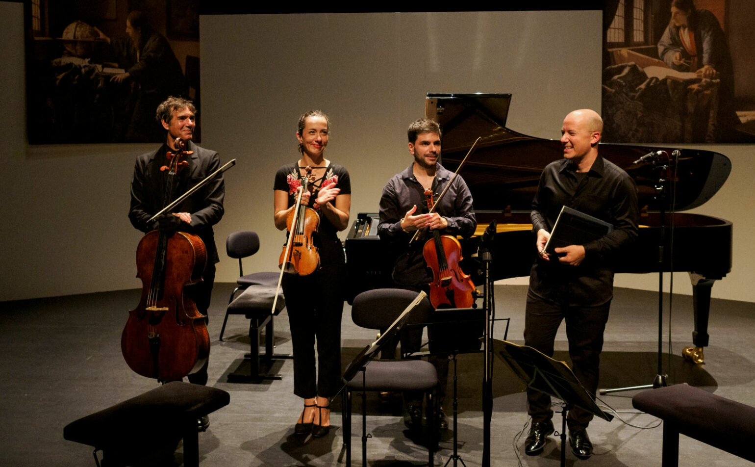Quatre musiciens, instruments à cordes et partitions, se tiennent sur scène devant un piano, semblant conclure une performance. Des peintures sont visibles sur le mur derrière eux.