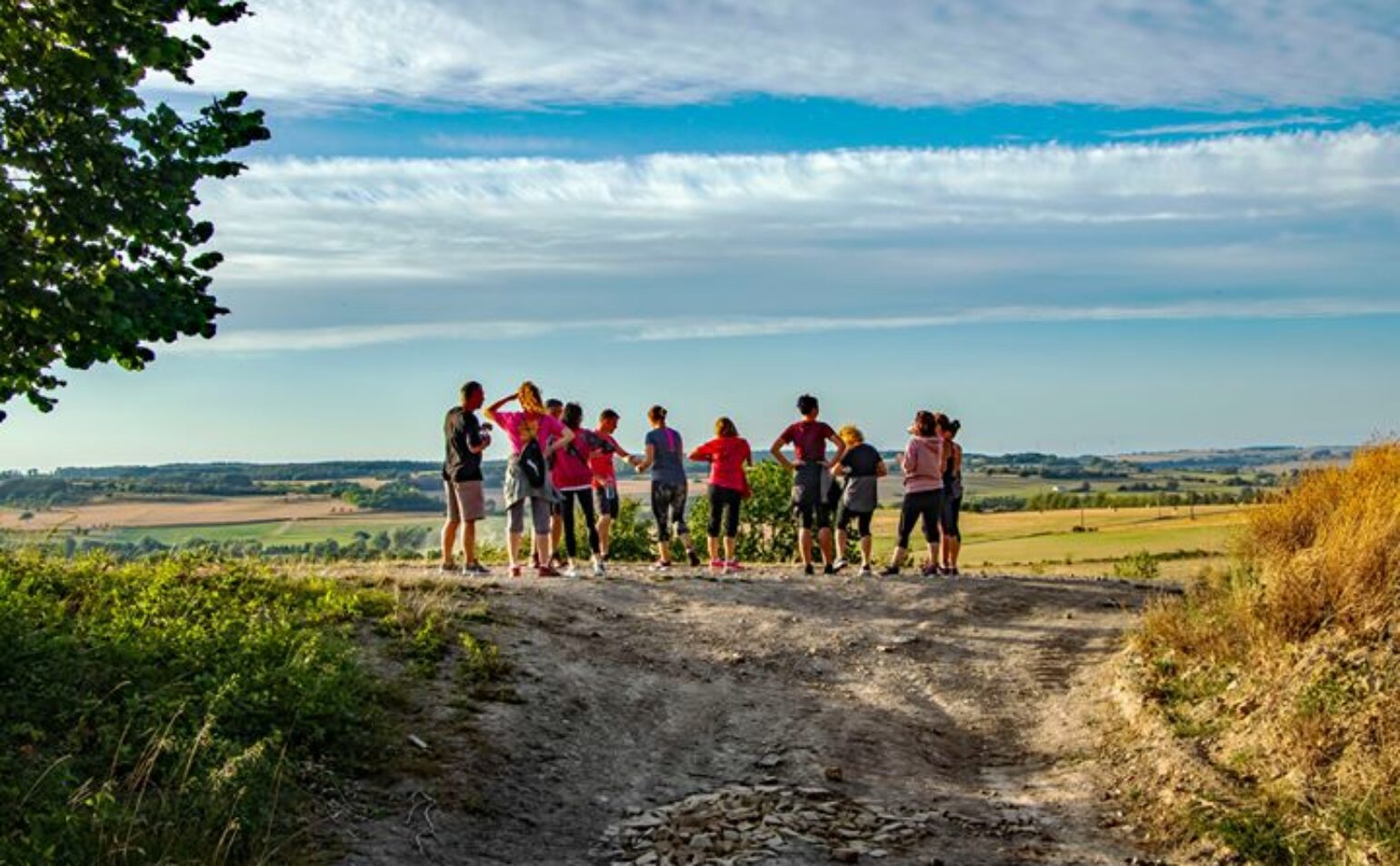 Un groupe de personnes se tient sur un chemin de terre surplombant des champs ouverts sous un ciel partiellement nuageux, certains faisant face au paysage et d'autres discutant entre eux.
